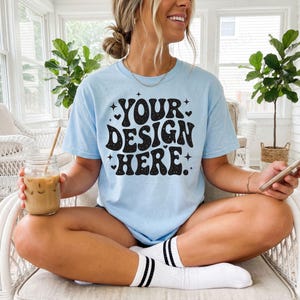 May include: Light blue t-shirt with the text "YOUR DESIGN HERE" in a black, retro font. The shirt is short-sleeved and has a crew neck. The person is wearing white socks with black stripes.