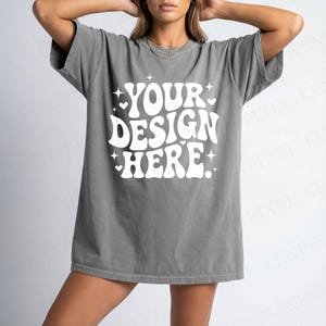 Puede incluir: Camiseta gris oversize con el texto "YOUR DESIGN HERE" en blanco. La camiseta tiene un corte holgado y mangas cortas. El texto est&aacute; en una fuente retro con estrellas y corazones.