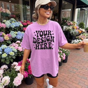 May include: A lilac-colored t-shirt with the text "YOUR DESIGN HERE" in black, with a retro font. The shirt is worn with black shorts and white sneakers. The background includes a flower shop with various colorful flowers.