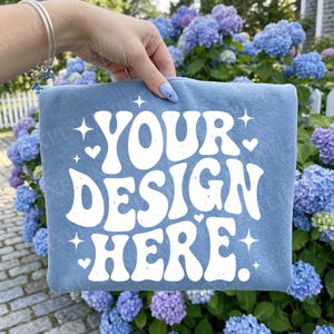 Puede incluir: Una bolsa azul claro con el texto blanco "YOUR DESIGN HERE", decorada con estrellas y corazones. La bolsa se muestra frente a un fondo de hortensias azules y moradas.