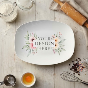 May include: White oval serving platter with floral design and text "YOUR DESIGN HERE". Baking ingredients like flour, eggs, and chocolate chips surround the platter. A rolling pin and whisk are also visible.