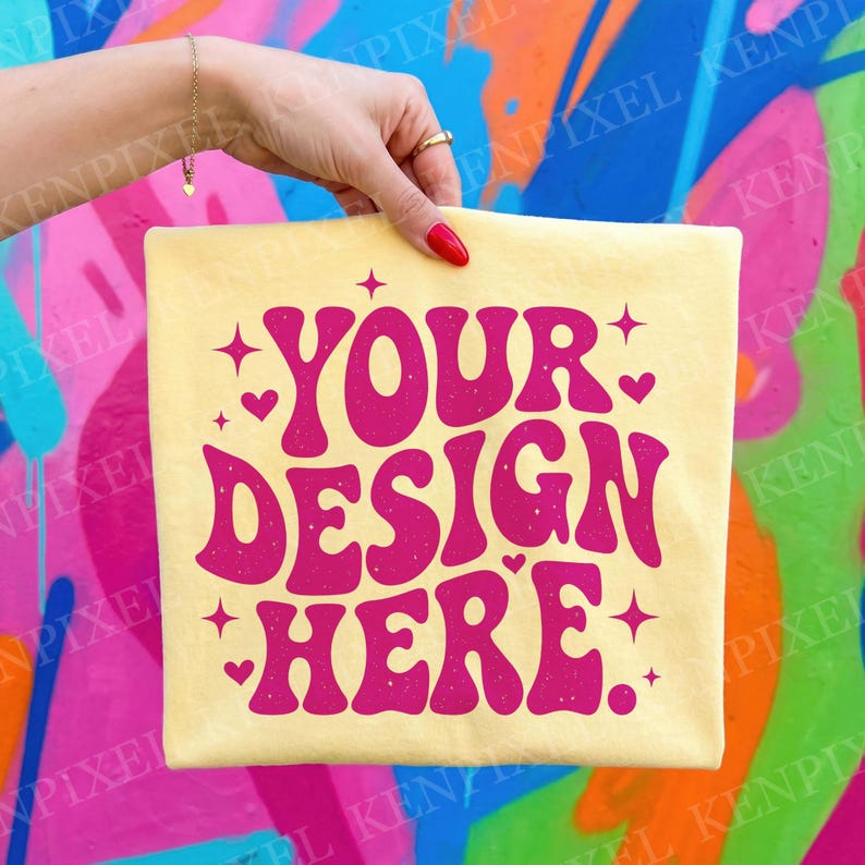 Peut inclure: Un t-shirt jaune clair avec le texte "YOUR DESIGN HERE" en rose, dans une police r&eacute;tro. Le motif comprend des &eacute;toiles et des c&oelig;urs. Le t-shirt est tenu par une main avec du vernis &agrave; ongles rouge.