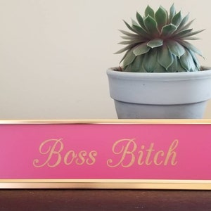 May include: Pink desk nameplate with gold trim and the text "Boss Bitch" in gold lettering.
