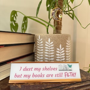 Könnte beinhalten: Ein Schreibtischschild mit dem Text "I dust my shelves but my books are still FILTHY" in rosa Schrift. Das Schild hat einen goldenen Rahmen und steht auf einer Holzoberfläche. Eine Topfpflanze und ein Stapel Bücher befinden sich im Hintergrund.
