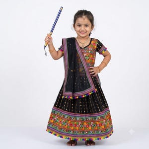 Könnte beinhalten: Ein schwarzes Lehenga-Choli-Set mit aufwendigen, mehrfarbigen Stickereien, Spiegelarbeiten und einem passenden Dupatta mit Bommelbesatz. Der Rock hat einen breiten gemusterten Saum und das Oberteil hat kurze Ärmel. Ein blau-goldener Stab wird gehalten.