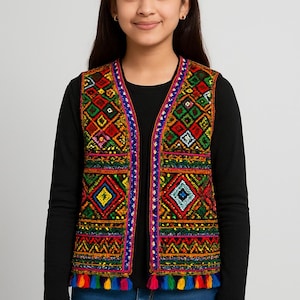 May include: A colorful, embroidered vest with a geometric pattern in red, yellow, green, and blue. The vest has a black lining and is trimmed with a blue border and colorful tassels at the hem. The vest is worn over a black long-sleeved shirt and blue jeans.