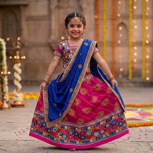 May include: A vibrant lehenga choli set featuring a bright pink skirt with gold peacock feather patterns and a wide, multicoloured embroidered border. The matching blouse has short sleeves and intricate mirror work, paired with a royal blue dupatta.
