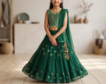 Puede incluir: Un lehenga verde, un atuendo tradicional indio, con una falda escalonada con adornos dorados y plateados. La parte superior a juego tiene un corpiño con lentejuelas y una bufanda drapeada. El atuendo lo lleva una persona.