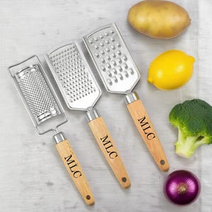 May include: Three stainless steel graters with wooden handles, each engraved with "MLC". A potato, lemon, broccoli, and red onion are arranged around the graters. The graters are designed for kitchen use.