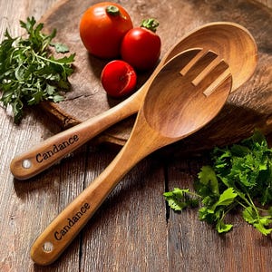 May include: Two wooden cooking spoons, one slotted, with the name "Candance" engraved on the handles. The spoons are on a wooden cutting board with tomatoes and parsley. The spoons are made of a light brown wood.