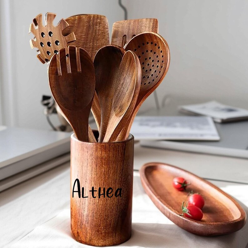 May include: A set of wooden kitchen utensils, including spoons, spatulas, and a slotted spoon, displayed in a wooden holder. The holder is engraved with the name "Althea". A small wooden serving dish with three red tomatoes is also present.