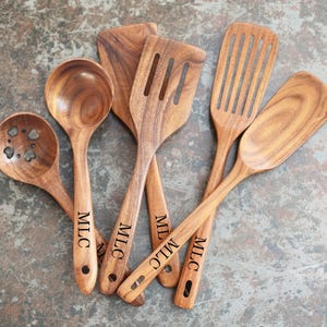 May include: A selection of wooden kitchen utensils, including a ladle, slotted spoon, and spatulas. The utensils are made of light brown wood and have the initials "MLC" engraved on the handles. The utensils are arranged on a textured surface.