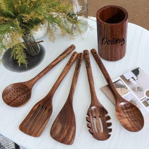 May include: A set of wooden kitchen utensils, including a slotted spoon and spatula. The dark brown utensils have the word "Destiny" engraved on the handles. A matching wooden utensil holder is also visible.