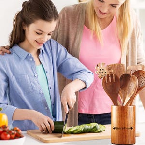 May include: A wooden utensil set in a wooden holder with the text "Baby Minna" is displayed. A person is slicing a cucumber on a wooden cutting board. Another person is standing next to them. The set includes various spoons and spatulas.