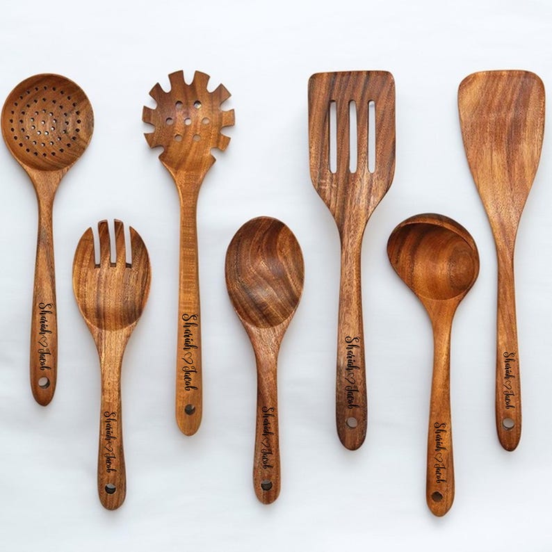 May include: A collection of seven wooden kitchen utensils, including a slotted spoon, pasta server, spatula, ladle, and serving spoons. Each utensil has a natural wood grain finish and the text "Shariah Jacob" engraved on the handle.