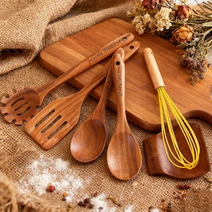 May include: A selection of wooden kitchen utensils, including a slotted spoon, spatula, and spoons, displayed on a wooden chopping board. A yellow whisk and a small wooden spatula are also present. The utensils are engraved with the name "JOHN SMITH".