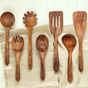 May include: A collection of seven wooden kitchen utensils, including a slotted spoon, pasta server, spatula, and serving spoon. Each utensil has a dark wood grain and the name "Shariah Jacob" engraved on the handle. They are arranged on a beige cloth.