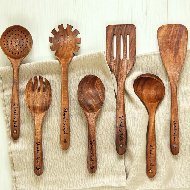 May include: A collection of seven wooden kitchen utensils, including a slotted spoon, pasta server, spatula, and serving spoon. Each utensil has a dark wood grain and the name "Shariah Jacob" engraved on the handle. They are arranged on a beige cloth.