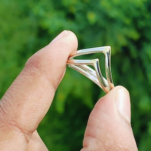 May include: A silver ring with a double chevron design. The ring is held between fingers against a blurred green background. The ring's geometric shape is modern and minimalist.