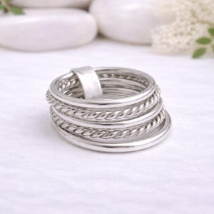 Op de afbeelding: Een stapel zilveren ringen met verschillende ontwerpen, waaronder gladde banden en gedraaide touwtexturen. De ringen worden gepresenteerd op een wit oppervlak, met witte stenen en bloemen op de achtergrond. Een eenvoudig en elegant sieraad.