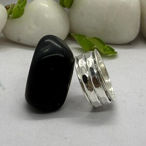 May include: A silver spinner ring with a hammered texture, next to a black stone. The ring has three bands, with the center band featuring a textured design. The background includes white stones and green leaves.