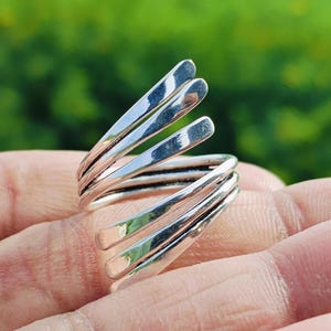 Könnte beinhalten: Ein silberfarbener Wickelring mit Spiraldesign. Der Ring hat vier gebogene, parallele Bänder, die sich um den Finger wickeln. Der Ring hat eine polierte, reflektierende Oberfläche, und der Hintergrund ist ein verschwommenes Grün.