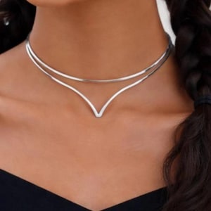 May include: A silver-colored choker necklace with a double-layered design. The top layer is a simple band, while the lower layer forms a V-shape at the center. The necklace is worn around the neck.
