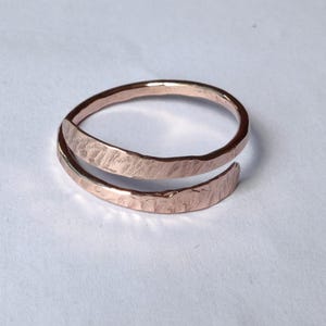 May include: A hammered copper ring with an open, wrap-around design. The ring features a textured surface and a warm, reddish-brown colour. The ring's band is approximately 0.6 cm wide.