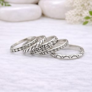 Może przedstawiać: Cztery srebrne pierścionki o różnych wzorach. Jeden pierścionek ma małe kamienie, a inne mają kwiatowe i wzorzyste detale. Pierścionki są na białym tle, co sugeruje motyw biżuterii lub akcesoriów.