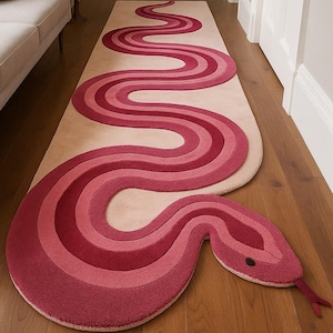 Puede incluir: Una alfombra decorativa con forma de serpiente, en tonos rosas y rojos. El diseño de la alfombra incluye un cuerpo sinuoso y una cabeza detallada con un ojo negro y una lengua roja. La alfombra mide aproximadamente 250 cm de largo.