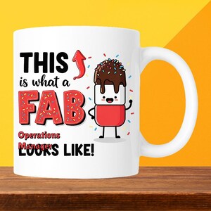 May include: White ceramic mug with a fun design. The mug has the text "THIS is what a FAB Operations Managers LOOKS LIKE!" in black and red, and a cartoon marshmallow character with chocolate and sprinkles.