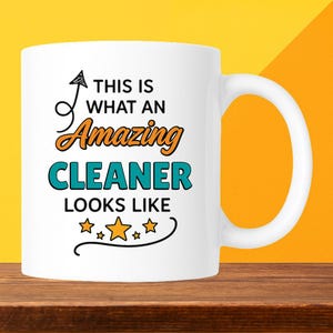 May include: White ceramic mug with a large handle. The mug features the text "THIS IS WHAT AN Amazing CLEANER LOOKS LIKE" in black, orange, and teal lettering, with star accents.