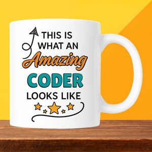 May include: White ceramic mug with the text "THIS IS WHAT AN Amazing CODER LOOKS LIKE" in black, orange, and teal. The mug has a curved handle and is decorated with stars. The mug is on a wooden surface.