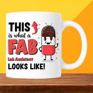 May include: White ceramic mug with the text "THIS is what a FAB Lab Assistant LOOKS LIKE!" in black and red. The word "FAB" is designed to look like a donut with sprinkles. A cartoon marshmallow character is also featured.