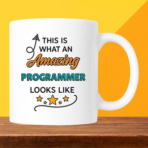 May include: White ceramic mug with the text "THIS IS WHAT AN Amazing PROGRAMMER LOOKS LIKE" in black, orange, and teal. The mug has a curved handle and is set against a yellow and brown background.