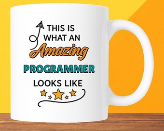 May include: White ceramic mug with the text "THIS IS WHAT AN Amazing PROGRAMMER LOOKS LIKE" in black, orange, and teal. The mug has a curved handle and is set against a yellow and brown background.