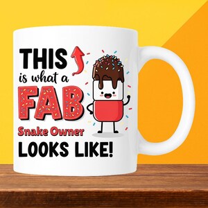 May include: White ceramic mug with a fun design. The mug has the text "THIS is what a FAB Snake Owner LOOKS LIKE!" in black and red, and a cartoon marshmallow character with chocolate frosting and sprinkles.