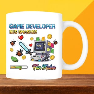 May include: White ceramic mug featuring a colourful graphic design. The design includes the text "GAME DEVELOPER BUG SMASHER" and "Fun Maker", along with pixelated hearts, coins, and a sword. A computer with a controller is also featured.