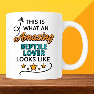 May include: White ceramic mug with the text "THIS IS WHAT AN Amazing REPTILE LOVER LOOKS LIKE" in black, orange, and teal. The mug has a curved handle and a black arrow design. The mug is on a wooden surface.