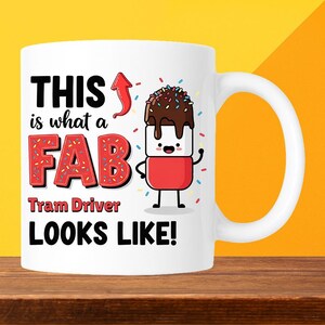 May include: White ceramic mug with a handle, featuring the text "THIS is what a FAB Tram Driver LOOKS LIKE!" in black and red. A cartoon marshmallow character with chocolate and sprinkles is also on the mug.