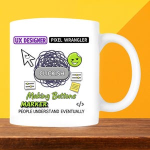 May include: White ceramic mug with a handle, featuring graphic design elements. The design includes a computer cursor, a smiley face sticker, and the text "UX DESIGNER PIXEL WRANGLER". Additional text reads "CLICKISH", "Making Buttons", and "PEOPLE UNDERSTAND EVENTUALLY".