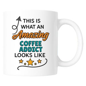 Op de afbeelding: Witte keramische koffiemok met een gebogen handvat. De mok heeft de tekst "THIS IS WHAT AN Amazing COFFEE ADDICT LOOKS LIKE" in zwarte, oranje en teal letters, met steraccenten.