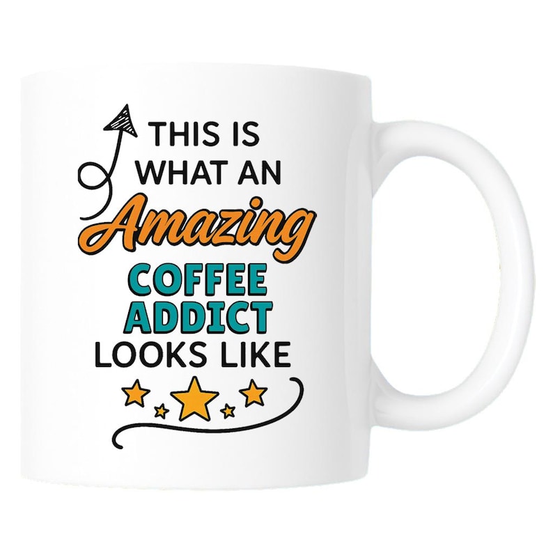 Op de afbeelding: Witte keramische koffiemok met een gebogen handvat. De mok heeft de tekst "THIS IS WHAT AN Amazing COFFEE ADDICT LOOKS LIKE" in zwarte, oranje en teal letters, met steraccenten.
