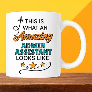 May include: White ceramic mug with a large handle. The mug features the text "THIS IS WHAT AN Amazing ADMIN ASSISTANT LOOKS LIKE" in black, orange, and teal lettering, with star accents. The mug is on a wooden surface with a yellow and orange background.