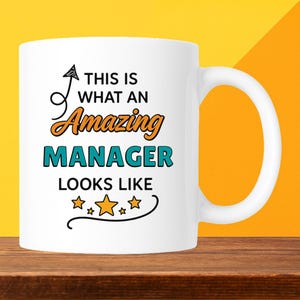 May include: White ceramic mug with the text "THIS IS WHAT AN Amazing MANAGER LOOKS LIKE" in black, orange, and teal. The mug has a curved handle and is set against a yellow and orange background.
