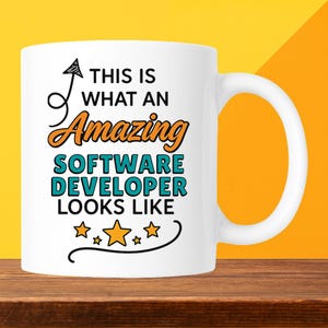 May include: White ceramic mug with the text "THIS IS WHAT AN Amazing SOFTWARE DEVELOPER LOOKS LIKE" in black, orange, and teal. The mug has a curved handle and is set against a yellow and orange background.