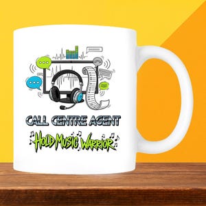 May include: White ceramic mug with a wraparound design featuring a headset, speech bubbles, and the text "CALL CENTRE AGENT Hold Music Warrior" in black and green. The mug is on a wooden surface with a yellow and orange background.