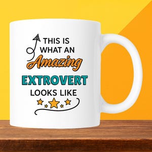 May include: White ceramic mug with the text "THIS IS WHAT AN Amazing EXTROVERT LOOKS LIKE" in black, orange, and teal. The mug has a curved handle and is set against a yellow and orange background.