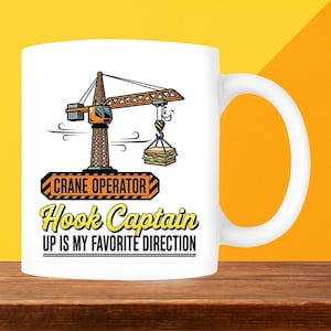 May include: White ceramic mug with a crane graphic and the text "CRANE OPERATOR Hook Captain UP IS MY FAVORITE DIRECTION". The crane is lifting a sandwich. The mug has a large, curved handle. The design is in orange and black.