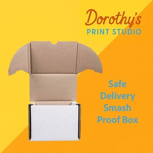 Op de afbeelding: Een open kartonnen doos met een witte binnenkant, tegen een gele en oranje achtergrond. De tekst "Dorothy's PRINT STUDIO" staat in de rechterbovenhoek, met "Safe Delivery Smash Proof Box" in blauwe tekst.